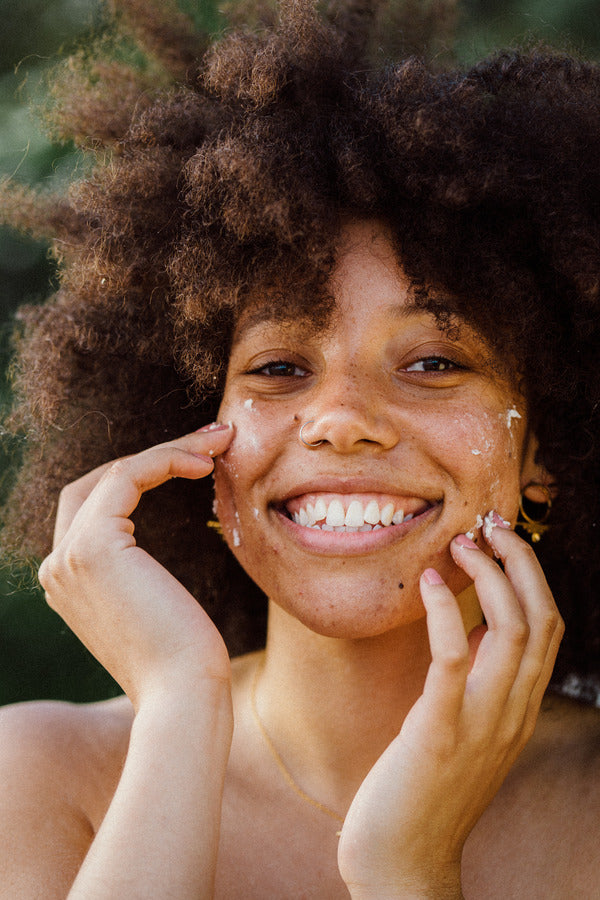 A young woman with an afro hairstyle is smiling brightly at the camera, her face dotted with Aviela pure shea butter. She is holding her hands up to her cheeks, gently massaging the product into her skin.