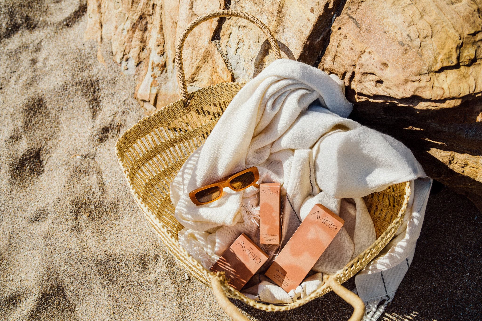 Natural, wholesome Aviela Skincare products: Shea Body Lotion, Shea hand butter & African Black Soap, in a woven basket with sun glasses on a beach