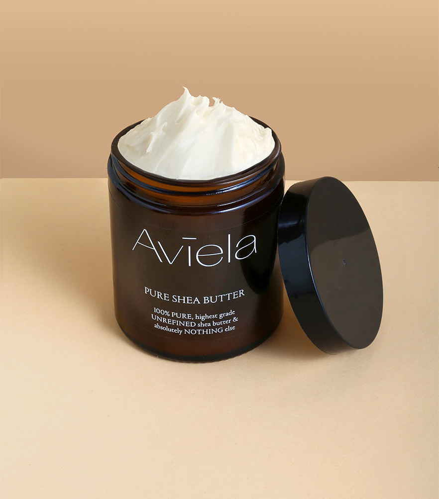A slightly elevated, close-up shot of an open jar of Aviela Pure Shea Butter. The dark amber glass jar is filled to the top with a whipped, creamy off-white 100% pure, highest grade unrefined shea butter that has peaks. The black lid is resting on the surface next to the jar. The background is a solid, light tan color.