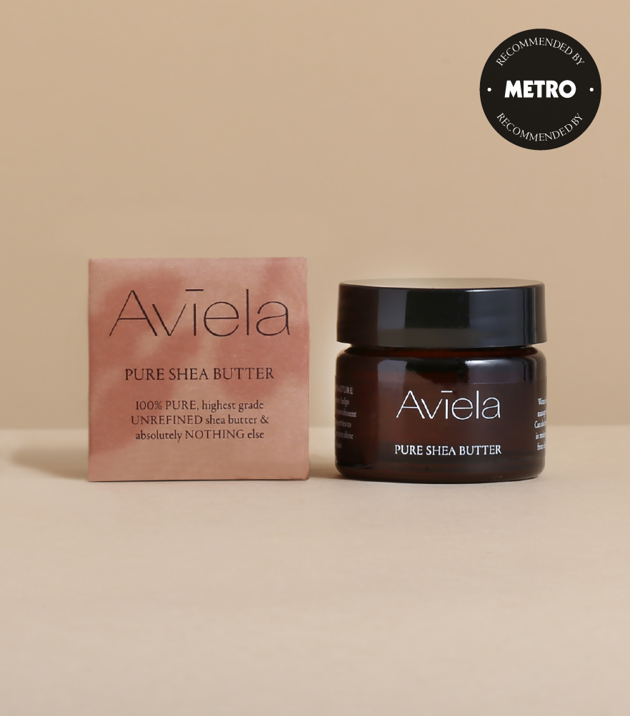A product shot featuring a small, square cardboard box and a dark amber glass jar of Aviela Pure Shea Butter next to it. Both the box and the jar have the same label design: the brand name "Aviela" in white text, followed by "PURE SHEA BUTTER" and "100% PURE, highest grade UNREFINED shea butter & absolutely NOTHING else". In the top right corner of the image is a circular badge that reads "RECOMMENDED BY METRO". The background is a solid tan color.