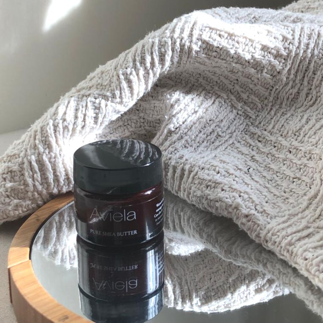 A jar of Aviela Pure Shea Butter with a black lid rests on a circular mirror, its reflection visible below. The jar has a dark amber color and a dark label with "Aviela" written in white. A textured, beige-colored knitted blanket is draped behind and to the right of the jar.