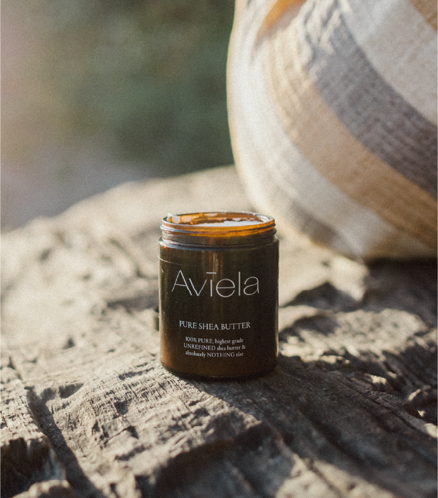 A jar of Aviela Pure Shea Butter with a black lid and a dark label sits on a weathered, light-colored wooden surface. The jar has a dark amber color. The label has the brand name, "Aviela" in white lettering at the top, followed by the text, "PURE SHEA BUTTER" and "100% PURE, highest grade UNREFINED shea butter & absolutely NOTHING else" below. A woven basket is partially visible in the blurred background.