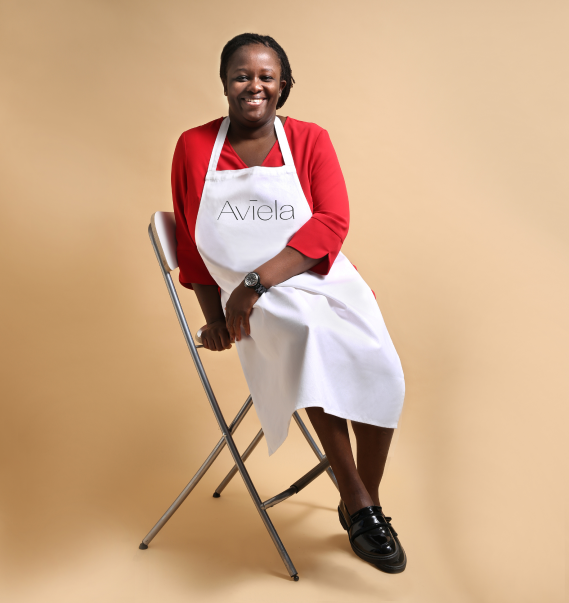 Aviela banner image of Patricia Monney, the founder of Aviela Skincare. She is seated on a white folding chair, smiling at the camera. They are wearing a red long-sleeved shirt and a white apron with the word "Aviela" in a simple font on the chest.