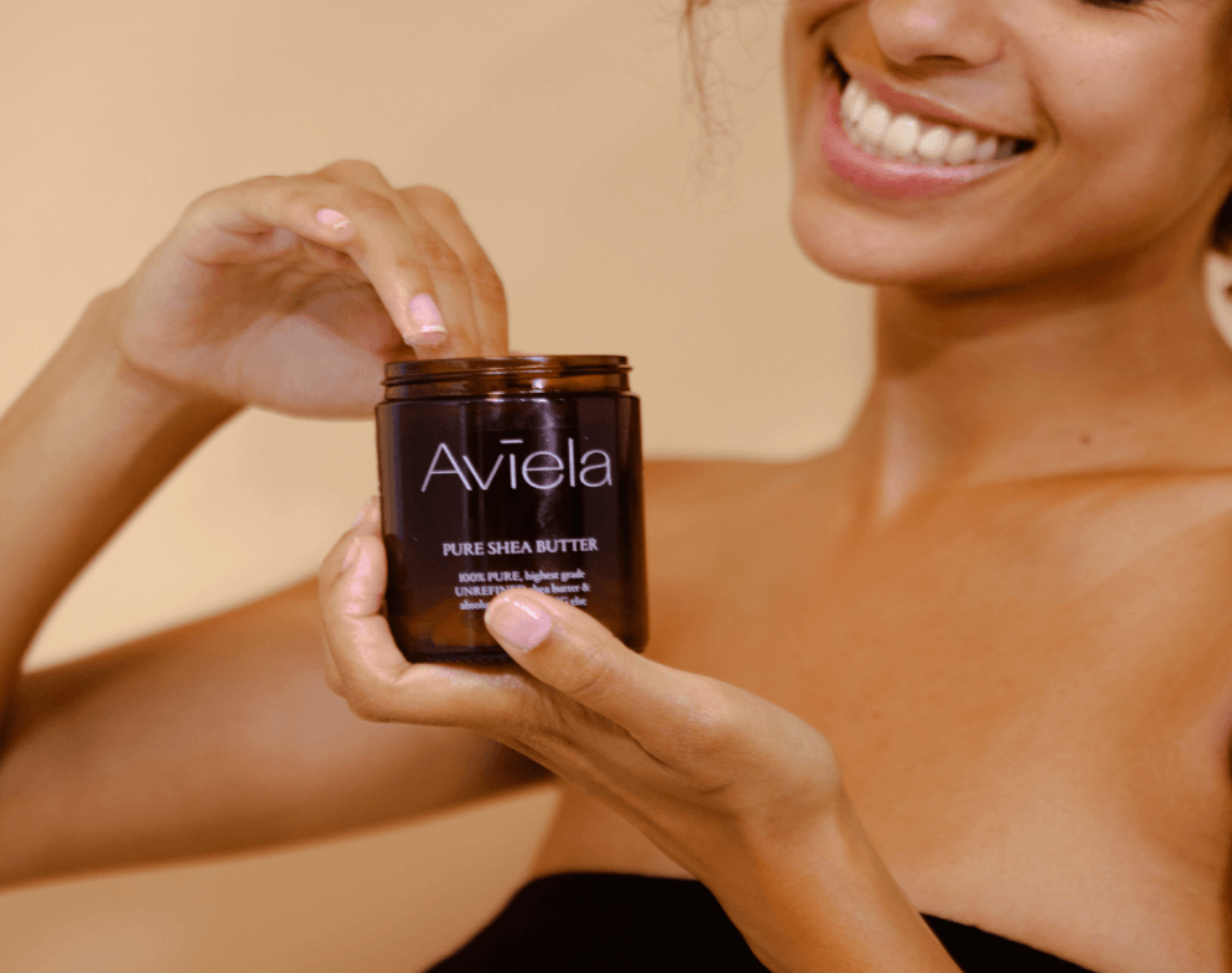 A smiling person holds a brown jar of Aviela Pure Shea Butter. The person's hands are visible as they dip their fingers to use the smooth shea butter from the jar, which has a label with light-colored text. The person is wearing a black top. The background is a solid, light color.