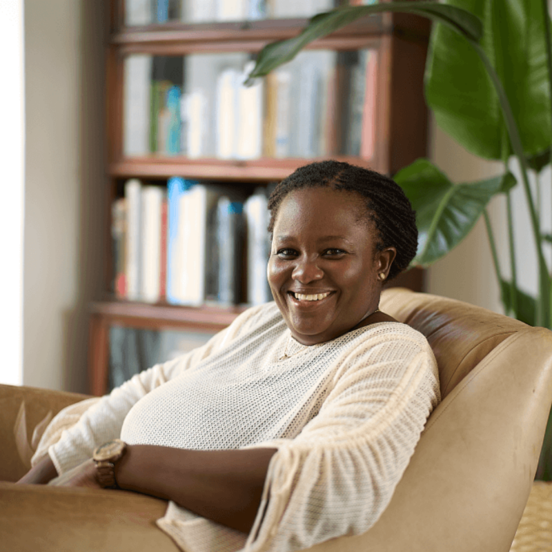 Patricia Monney, founder of Aviela Skincare with her hair in braids is seated in a light brown armchair, smiling at the camera. She is wearing a cream-colored, long-sleeved top and a watch. A bookshelf is visible in the background, along with a large green plant.
