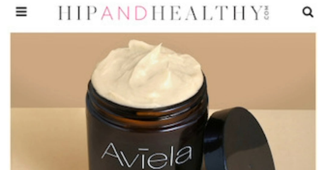 A screenshot of a webpage from "hipandhealthy.com" featuring a jar of Aviela product Pure Shea Butter. The jar is dark brown with a black lid resting next to it. The jar is open, revealing a generous amount of thick, white cream or butter with a peak on top. The product name "Aviela" is visible on the side of the jar.