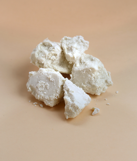 A banner image of chunks of raw, unrefined shea butter is shown on a solid tan background. The shea butter has a rough, somewhat flaky texture and a light, off-white to yellowish color sourced for Aviela Skincare.