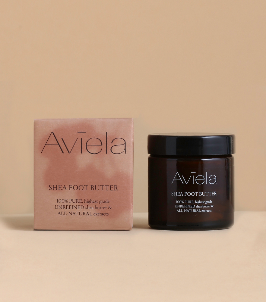 A jar and its corresponding box for Aviela Shea Foot Butter are placed side-by-side on a light-colored surface. The jar is dark brown with a black lid, and its label has light-colored text. The rectangular box is a peach color with a light, watercolor-like pattern. Both the jar and the box have the product name and a description of the ingredients.